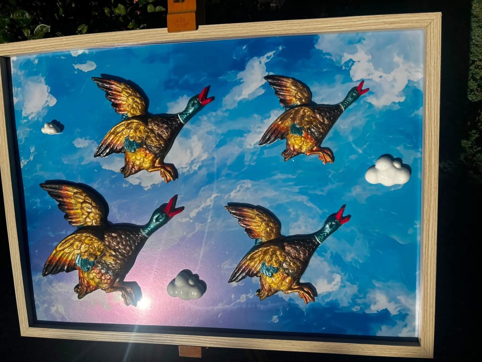Four bronze and gold ducks mounted in a wooden box frame against a blue sky with matching clouds