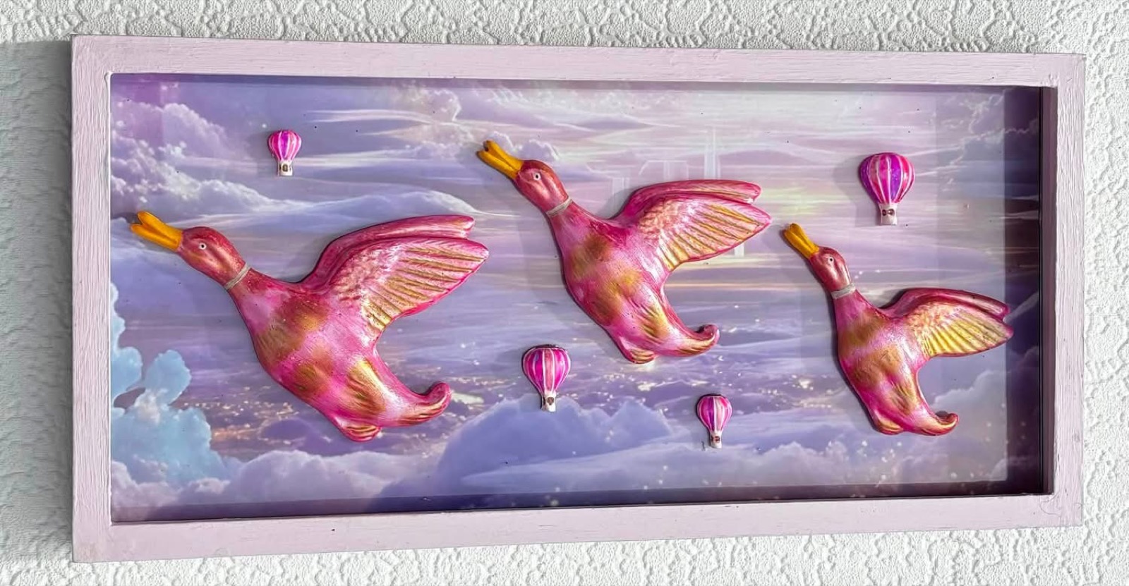 Three pink and gold ducks mounted in a pink box frame against a painted lilac sky with small hot air balloons